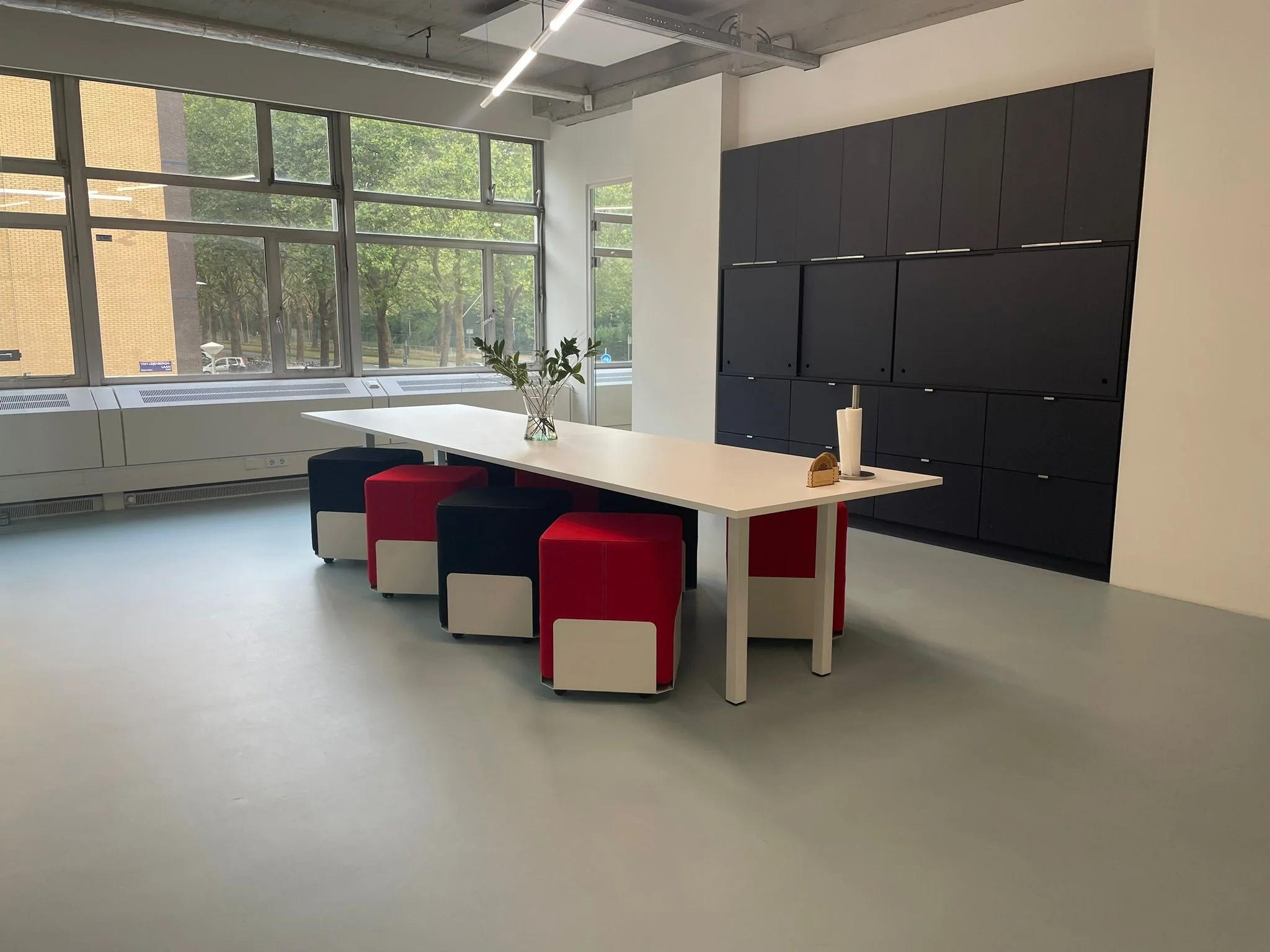 Moderne vergaderruimte aan de Van Leijenberghlaan met een lange tafel, kleurrijke zitblokken en grote ramen met uitzicht op bomen.